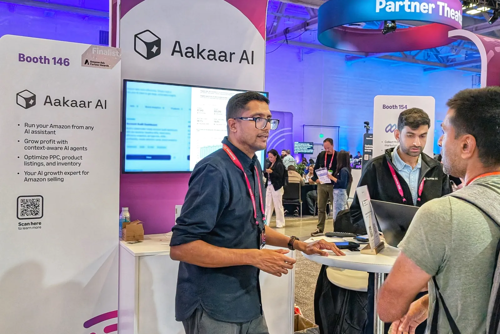 Man in a dark shirt and lanyard talking with a visitor at the Aakaar AI booth, Booth 146, beside a screen showing a dashboard
