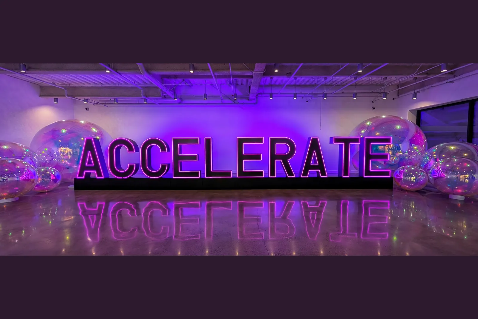 Large illuminated ACCELERATE letters in a purple-lit room, reflected in a glossy floor, with iridescent balloons on both sides